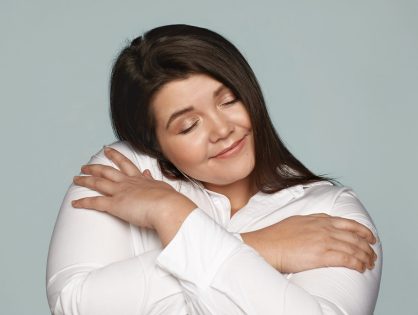 Self love and appreciation concept. Picture of beautiful charming young plus size female with large curvy body embracing herself, keeping hands on her shoulders, closing eyes and smiling happily